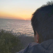 Jay-Ar Foronda, a young Filipino American man with short black hair, leaning against a wooden wall by the oceanside watching a beautiful sunset on a shore of the beach.